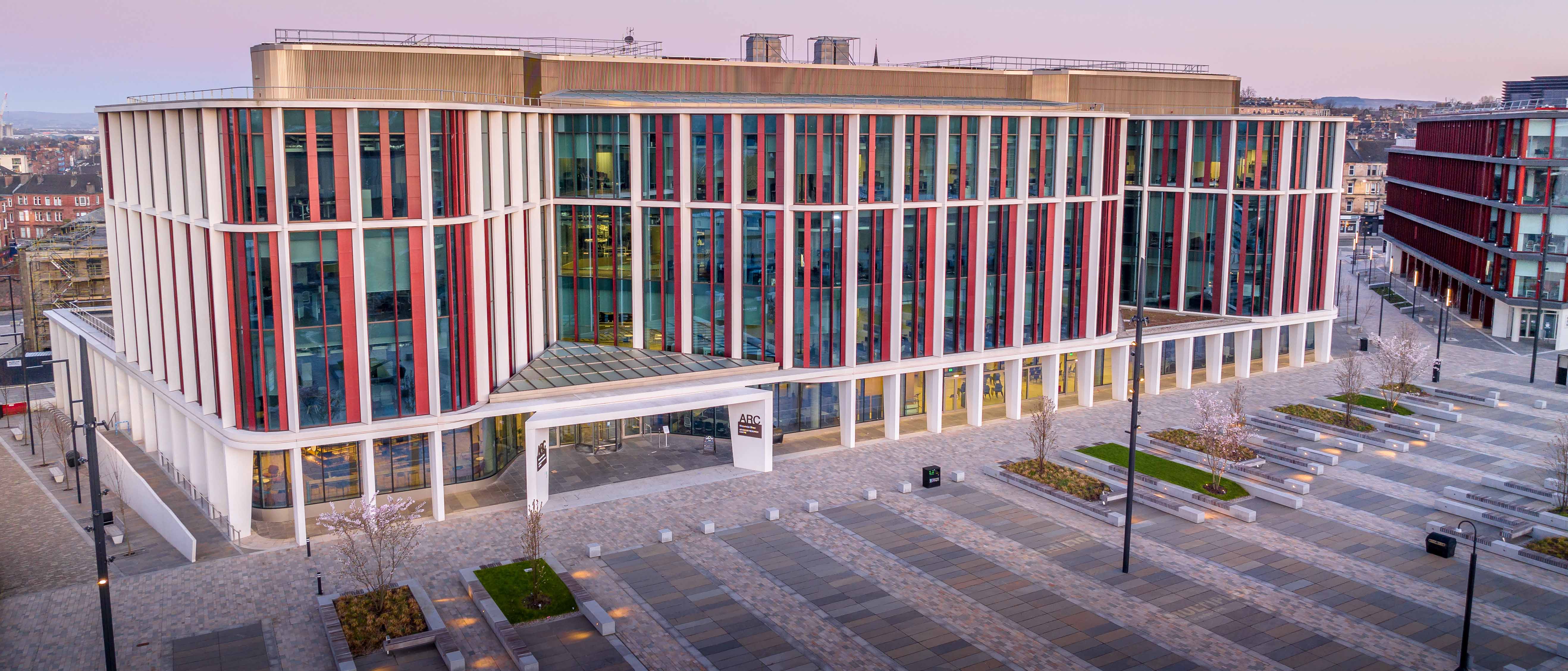 A picture of a large modern building, mostly glass-fronted with white framing and red accents behind an open square with trees and seating spaces.