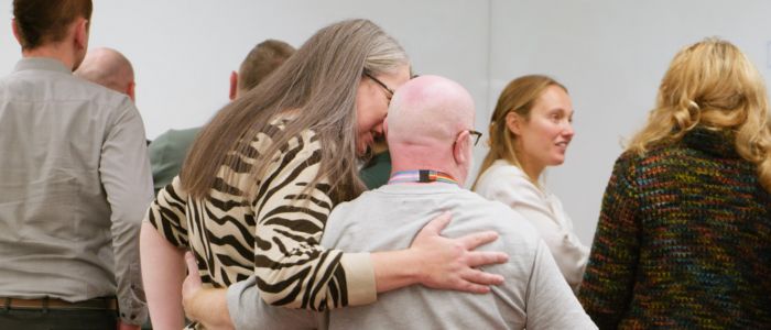 Two individuals in the foreground, one wearing a zebra-striped sweater and the other with glasses and a lanyard displaying LGBTQIA flags, engaged in conversation with an arm around the other, surrounded by others interacting.