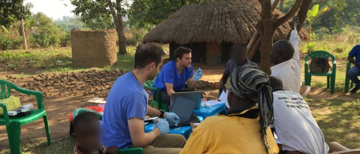 Healthcare workers in blue scrubs conducting a medical outreach in a rural outdoor setting, seated at a table with laptops and supplies, engaging with community members near green chairs and thatched-roof huts.