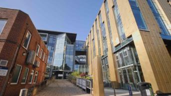 A wide-angle view of two modern CVR buildings, featuring glass façades and gold cladding, with a walkway and planted area leading towards a connecting glass bridge under a clear blue sky.