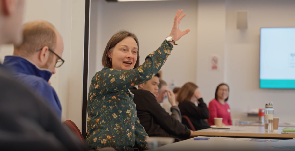 A person seated at a conference table raises their hand to contribute during a meeting. Several other attendees sit around the room, some listening and others taking notes.