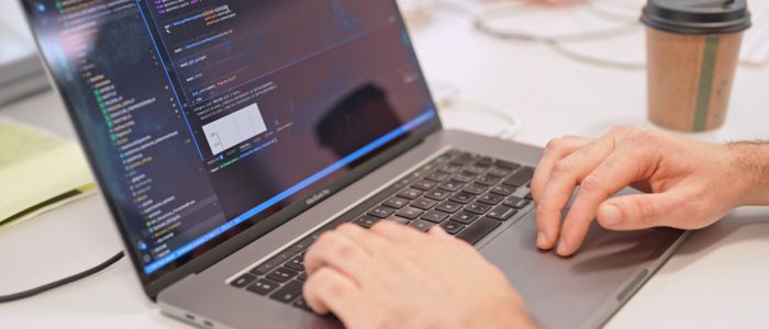 A person typing on a laptop displaying code in a development environment, with a coffee cup and documents on the desk nearby.