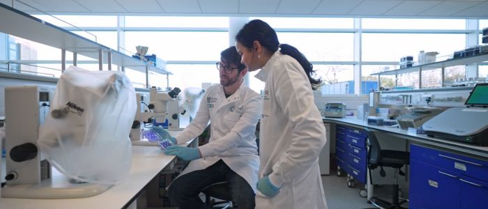 Two people in lab coats work together at a laboratory bench, examining a tray of samples beside microscopes and other equipment.
