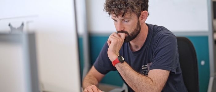 A person with short, curly hair sits at a desk in an office environment, resting their head on one hand while looking toward a computer screen. The person is wearing a dark short‑sleeved shirt and a red wristwatch. The background includes a whiteboard, partition panels.