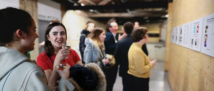 photo of people in front of a plywood wall with posters. in foreground two white women drink white wine