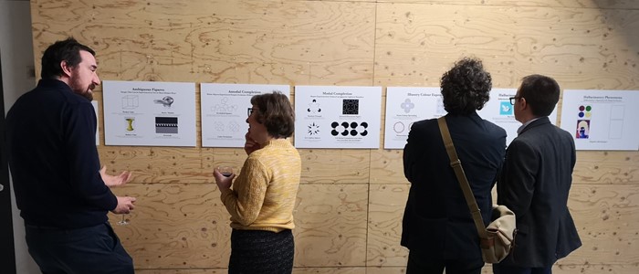 photo of a smaller white woman explaining something to a taller white man in front of a plywood wall with posters, with two other people on the right turned with their back to the camera
