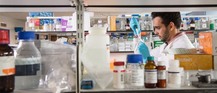 A man is using a pipette in a lab and is visible between shelves filled with bottle of chemicals and lab equipment.